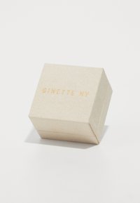 Scatola beige chiaro con superficie texture, leggermente inclinata; presenta la scritta in rilievo dorato "GINETTE NY" sulla parte superiore. Forma quadrata.