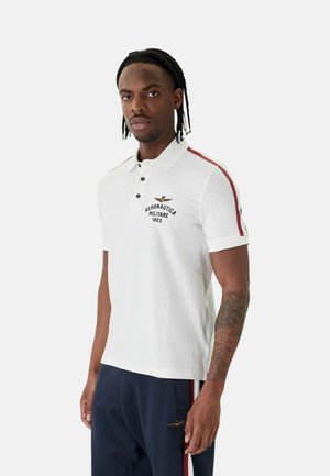 Uomo con capelli intrecciati che indossa una polo bianca con strisce rosse e il logo "Aeronautica Militare 1923", e pantaloni blu navy con una striscia abbinata.