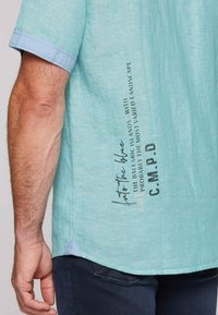 Lichtblauwe shirt met korte mouwen, gemaakt van een gestructureerde stof; heeft donkere tekst en accenten aan de zijkant en op de mouweinden. De zoom is gebogen.