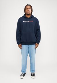 Felpa blu navy con tasca a marsupio frontale e coulisse, con il logo "JACK & JONES" bianco e rosso. Indossata con jeans azzurri chiari e sneakers nere.