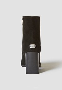 Bottine en suède noire avec un talon épais, fermeture éclair sur le côté et accent logo métallique à l'arrière. Texture lisse et design épuré.