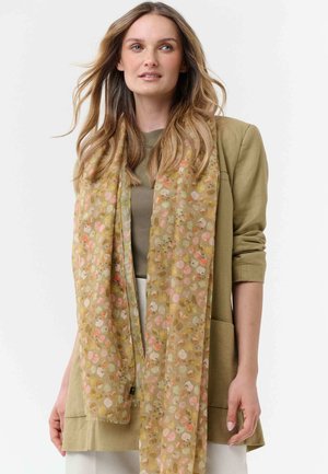 Frau mit langen, welligen Haaren, die einen olivgrünen Blazer und ein T-Shirt, weiße Hosen und einen hellen, floral gemusterten Schal trägt.