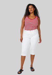 Rød og hvidstribet tanktop, hvide knælange bukser, sorte sandaler med nitter. Krøllet hår, stående stilling foran en grå baggrund.