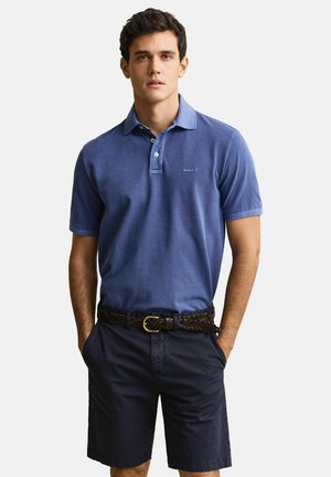 Mann trägt ein blaues Poloshirt und dunkelblaue Shorts mit geflochtenem braunem Gürtel, steht mit den Händen in den Taschen vor einem einfarbigen Hintergrund.