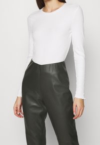 Weißes, geripptes Langarmoberteil, kombiniert mit dunkelgrünen, hochtaillierten, glänzenden Kunstlederhosen, die eine glatte Silhouette erzeugen.