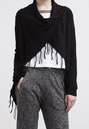 Osoba ubrana w czarny szal z frędzlami na białej bluzce i szare spodnie o fakturze, z rękami w kieszeniach na jednolitym tle.