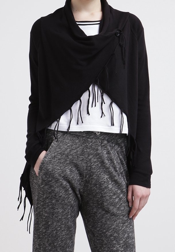 Personne portant un châle noir à franges sur un haut blanc et un pantalon gris texturé, les mains dans les poches, devant un fond uni.