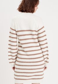 Robe en tricot avec des rayures verticales blanches et beiges. Elle présente un col montant, des manches longues et une texture côtelée, arrivant à mi-cuisse.