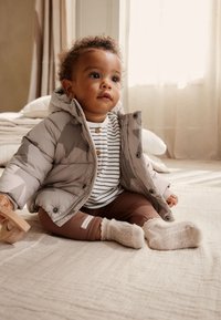 Veste matelassée camouflage grise avec capuche, chemise rayée blanche et marine, leggings bruns côtelés, et chaussettes en tricot crème. Jouet en bois tenu dans la main.