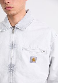 Denimjakke i lys grå med lynlås foran, krave og lomme med en gul logo patch. Struktureret stof med et slidt udseende.