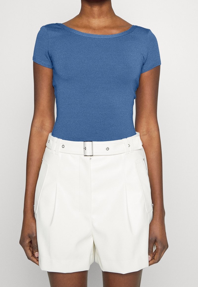 Personne portant un haut bleu ajusté à manches courtes et un short blanc taille haute avec ceinture et poches latérales zippées.
