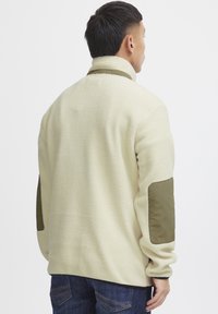 Fleece jacket in light cream with olive green elbow patches, features a high collar and relaxed fit. Soft texture and casual design.