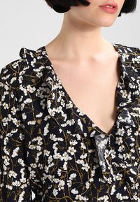 Abito a fiori con scollatura a V profonda e colletto a balze in blu navy, caratterizzato da fiori bianchi e finiture dorate su un tessuto morbido.