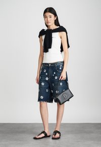 Débardeur blanc ajusté, shorts en denim foncé avec broderies florales, sac à main noir, sandales plates et un pull marine drapé sur les épaules.