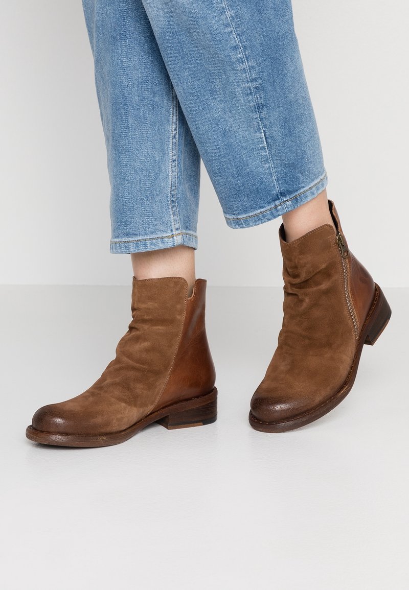 Bottines marron en daim et cuir avec fermeture éclair latérale, portées avec un jean en denim bleu clair coupé au-dessus de la cheville sur un fond blanc uni.