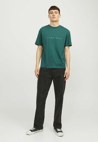 Dunkelgrünes Baumwoll-T-Shirt mit kurzen Ärmeln und Rundhalsausschnitt, versehen mit einem weißen Textlogo, kombiniert mit schwarzen Jeans und schwarzen Sneakers.