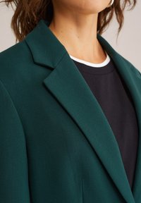 Zelený blazer s klopovým golierom, vyrobený z hladkej tkaniny. Nosí sa cez čiernu košeľu s bielym lemovaním. Detailný záber ukazuje textúru a detaily.