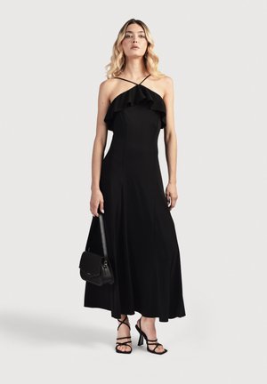 Mujer con un vestido midi de cuello halter negro con detalle de volantes, sosteniendo un bolso negro y llevando sandalias negras de tacón con tiras.