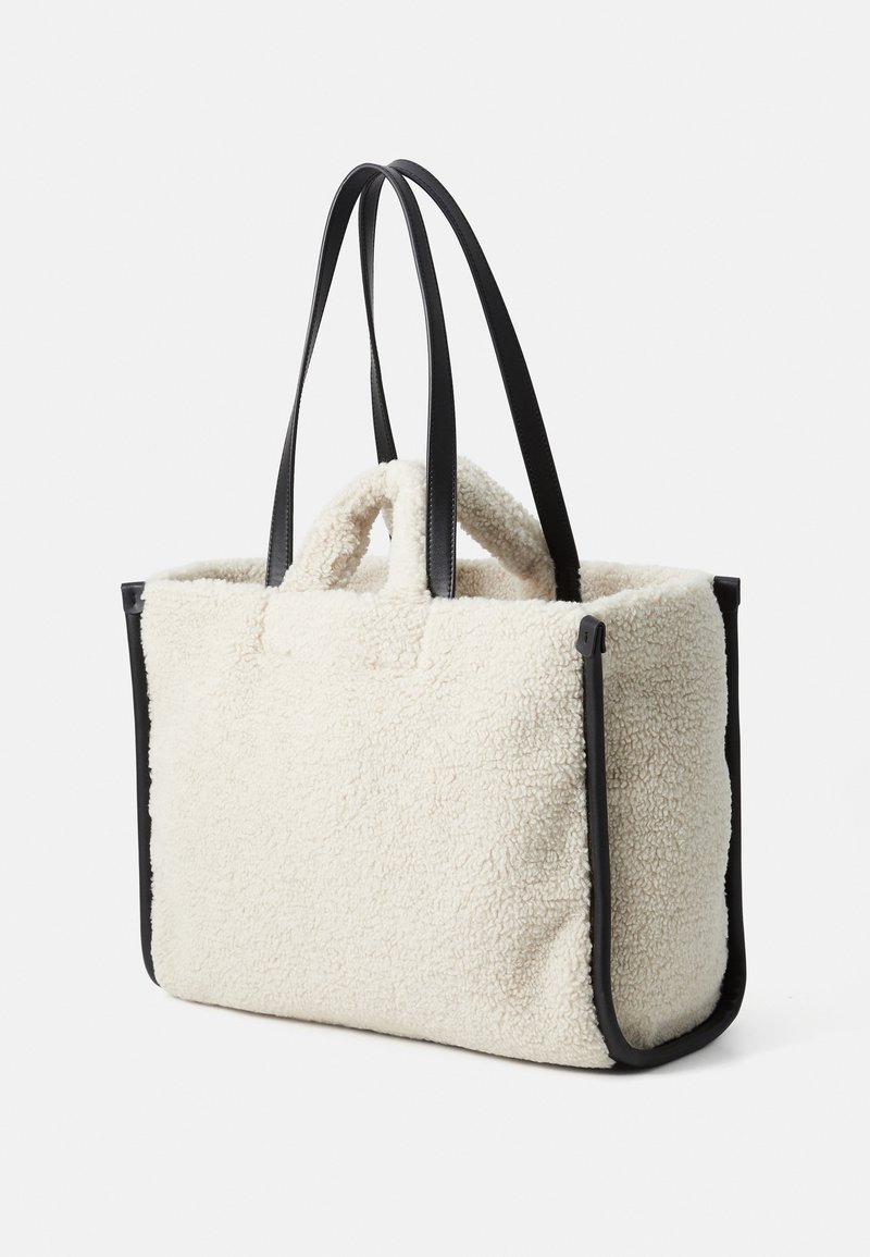 Cremefarvet tote taske lavet af fluffy, tekstureret materiale, med sorte læderdetaljer og lange, flade skulderstropper. To korte håndtag øverst.