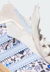 Helleblaue Sneaker mit einem gemusterten Obermaterial aus Stoff mit floralen Details, Wildlederakzenten und strukturiertem Overlay. Weiße Schnürsenkel und ein sichtbares Logoetikett.