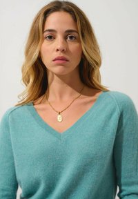 Suéter azul claro de cuello en V en material suave, combinado con un collar dorado que presenta un colgante redondo. El fondo es de color blanco liso.