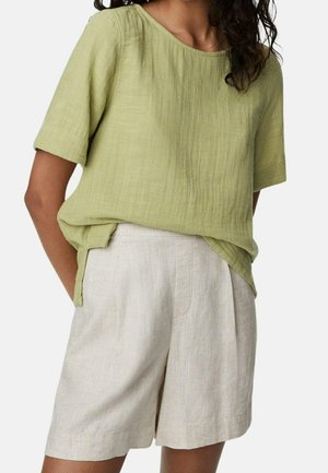 Frau trägt eine hellgrüne kurzärmelige, strukturierte Bluse, die in hoch taillierte beige Leinen-Shorts gesteckt ist, steht mit den Händen in den Taschen.