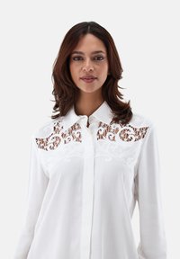 Camicia bianca con bottoni, caratterizzata da un design ricamato e ritagliato sulle spalle, con motivi floreali e una trama di tessuto liscia e leggera.