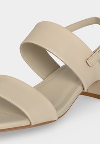 Sandale beige à bout ouvert avec larges brides sur le pied et la cheville, avec un talon bas et un matériau synthétique lisse.