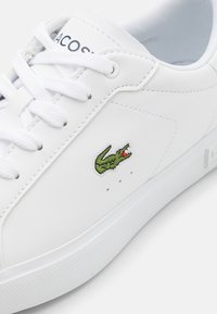 Vit lädersneaker med snörningsdesign. Har grönt broderat krokodil-logotyp, perforeringar för ventilation och en texturerad gummisula.