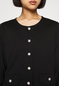 Černý cardigan s kulatým výstřihem, šesti dekorativními stříbrnými knoflíky a dvěma předními kapsami. Materiál vypadá hladce a má texturu.