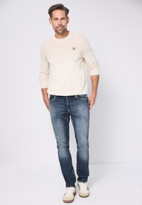 Svetle béžový pletený svetr s žebrovanou texturou a čtvercovým logem na hrudi, kombinovaný s tmavě modrými úzkými džínami a bílými teniskami.