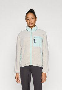 Ζακέτα από φλις σε ανοιχτό γκρι με λεπτομέρειες σε μέντα πράσινο. Διαθέτει τσέπη με φερμουάρ, ψηλό γιακά και ελαστικά μανίκια. Λεπτή υφή και χαλαρή εφαρμογή.