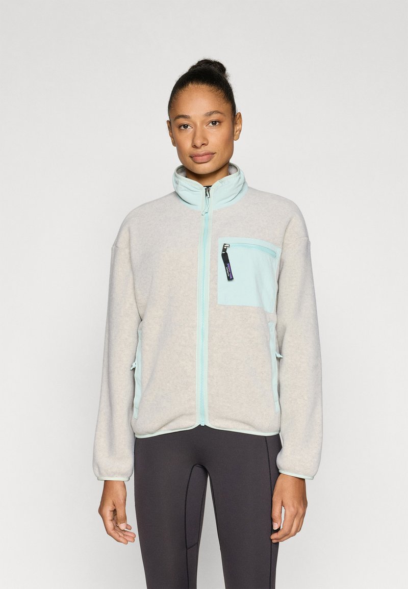 Ζακέτα από φλις σε ανοιχτό γκρι με λεπτομέρειες σε μέντα πράσινο. Διαθέτει τσέπη με φερμουάρ, ψηλό γιακά και ελαστικά μανίκια. Λεπτή υφή και χαλαρή εφαρμογή.