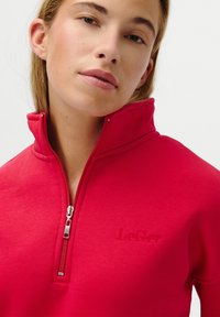 Pulover roșu cu fermoar jumătate, confecționat dintr-un material moale, cu un logo subtil embosat și un fermoar elegant, având o textură netedă pe întreaga suprafață.