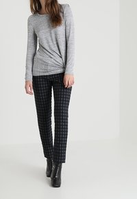 Kvinna som bär en ljusgrå långärmad tröja, svart och mörkblå rutiga byxor samt svarta ankelstövlar, stående framför en enfärgad bakgrund.