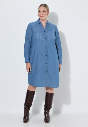 Kvinde iført en blå denim kjole med knapper og blomsterbroderi, kombineret med mørkebrune knæhøje støvler, stående mod en hvid baggrund.