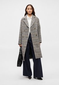 Διπλωμένος καρό παλτό σε γκρι, μαύρο και μπεζ, με μεγάλες πέτες και τσέπες. Φορεμένο πάνω από λευκό πουκάμισο και σκούρα παντελόνια με φαρδιά μπατζάκια.