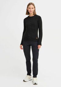 Svart kabelstickad tröja med långa ärmar, ribbad manschett och nederkant, med en liten logotypaccent på nedre vänstra sidan. Kombinerad med mörka byxor och sneakers.