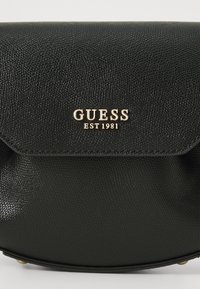 Svart läder crossbody-väska med texturerad yta, guld "GUESS EST 1981"-logotyp och en klaffstängning. Rundad form med guldaccenter.