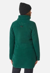 Cappotto in pile verde con colletto alto, tessuto testurizzato e logo sul retro. Presenta un orlo curvato e un anello per appenderlo. Indossato un berretto nero.