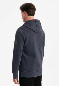 Felpa navy con cappuccio e cordino, foderata in tessuto a quadri, con polsini e orlo a coste, e una patch con logo sulla manica sinistra. Texture morbida.