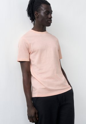 Moški s temno poltjo in dredljami ima na sebi svetloroza majico s kratkimi rokavi in črne hlače, stoji pred enobarvnim belim ozadjem.