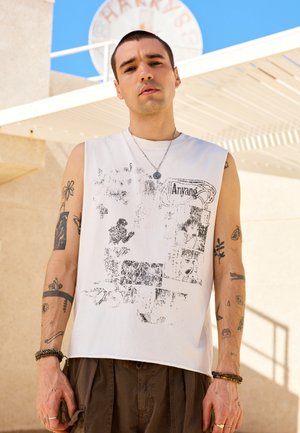 Giovane uomo con capelli corti e tatuaggi indossa una maglietta bianca senza maniche con grafica e pantaloni marroni, in piedi all'aperto sotto una tettoia bianca.
