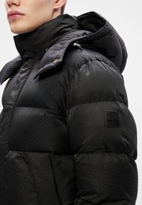 Μαύρο μπουφάν με γυαλιστερή υφή, διαθέτει αποσπώμενη κουκούλα, μοτίβο στο ύφασμα και μαύρο λογότυπο στη μανσέτα.