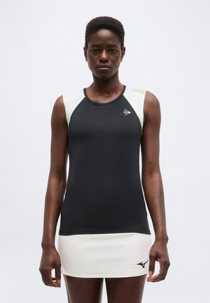 Mujer con una camiseta deportiva negra sin mangas con hombros blancos y verdes y una falda de tenis blanca, de pie contra un fondo liso.