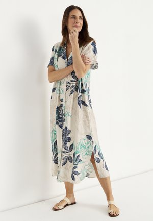 Kobieta stoi przy białej ścianie, nosi beżową sukienkę midi z niebiesko-zielonym kwiatowym wzorem i beżowe sandały z odkrytymi palcami.