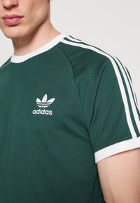 Žalias medvilninis marškinėliai su balta apykakle, baltais rankovių kraštais ir trimis baltais dryžiais ant pečių. Ant krūtinės pavaizduotas Adidas logotipas.