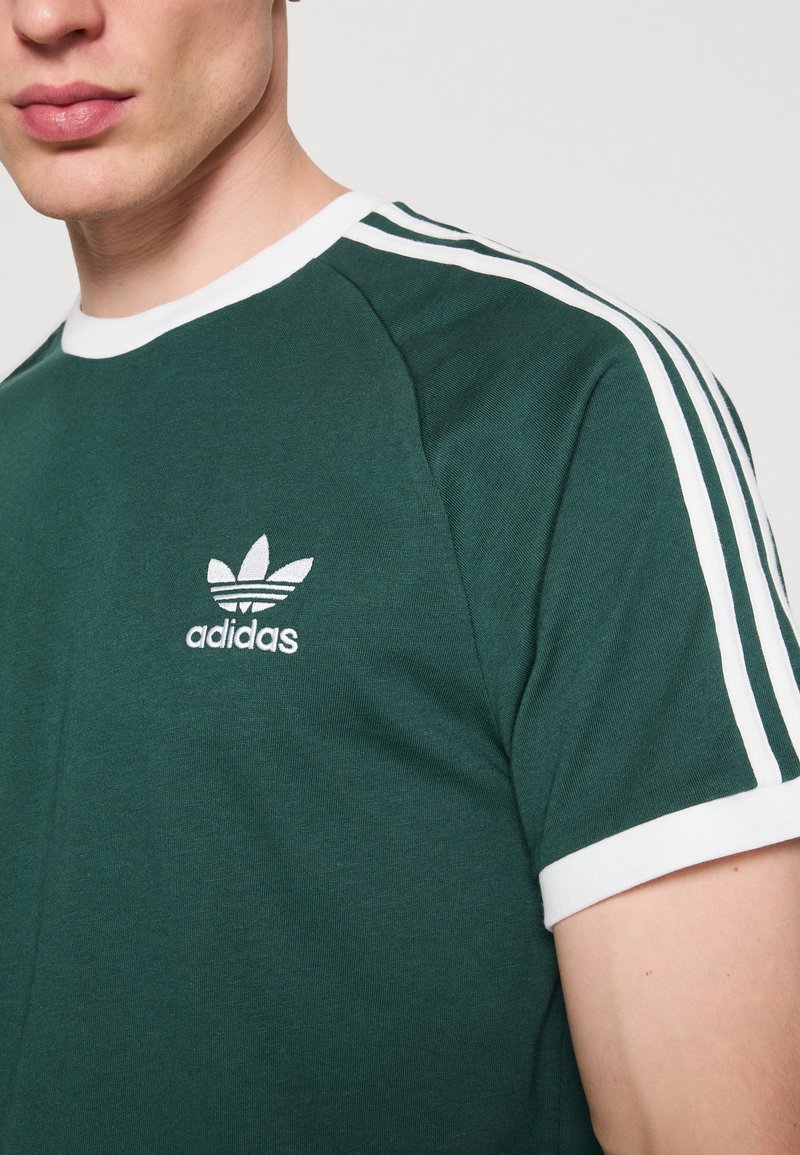 T-shirt en coton vert avec un col rond blanc, des bords de manches blancs et trois rayures blanches sur les épaules. Logo Adidas affiché sur la poitrine.