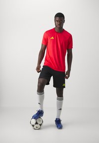 Rode sportt-shirt met gele accenten, zwarte shorts, witte sokken met zwarte strepen, blauwe voetbalschoenen en een voetbal.