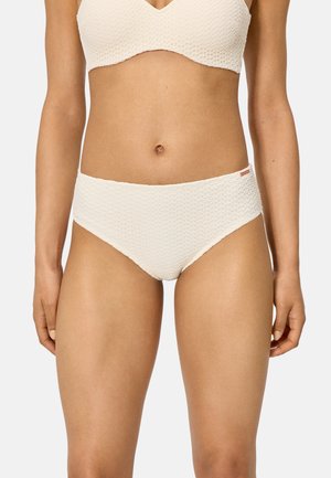 Mujer con bikini de cintura alta en tono marfil texturizado y top a juego, de pie con los brazos relajados sobre un fondo liso.